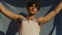 Cosimo Miccolis stands outdoors in warm sunlight with his arms extended, wearing a white tank top and briefs against draped fabric.