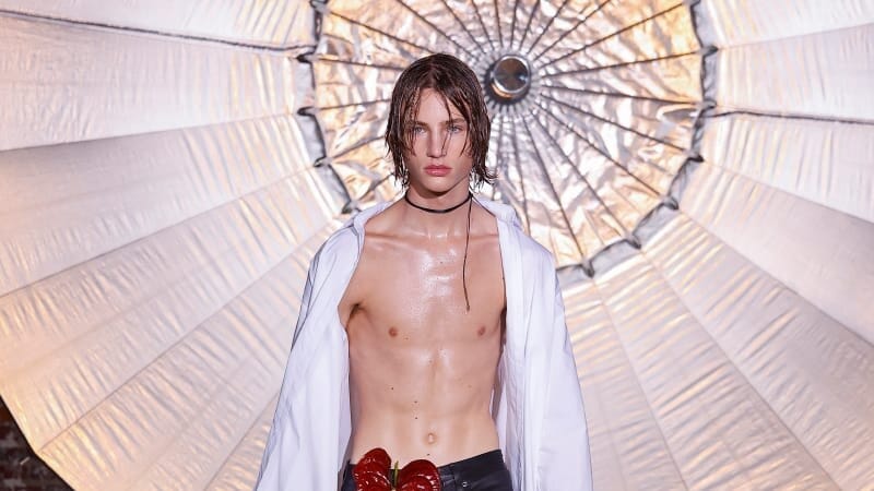 Runway model wears an open white shirt, black leather trousers, tall lace-up boots and a red floral accessory at the waist beneath a large illuminated structure.