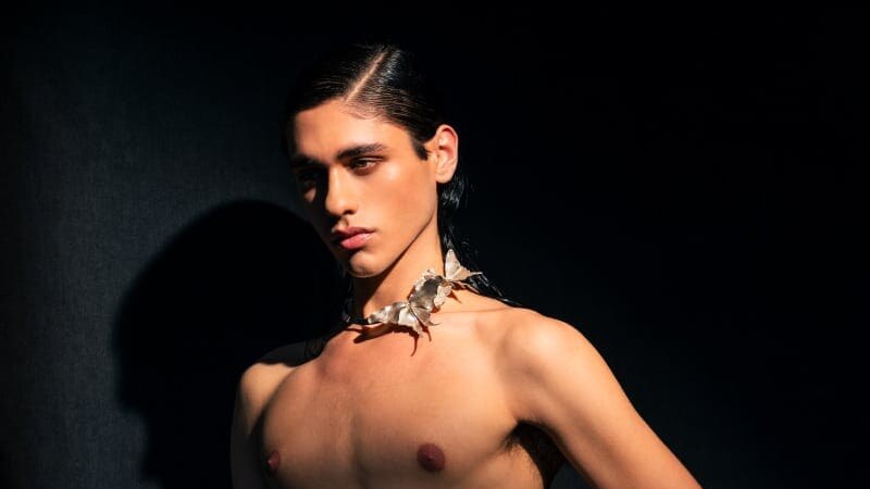 A shirtless model poses against a dark background wearing sculptural silver jewelry at the neck and hand.