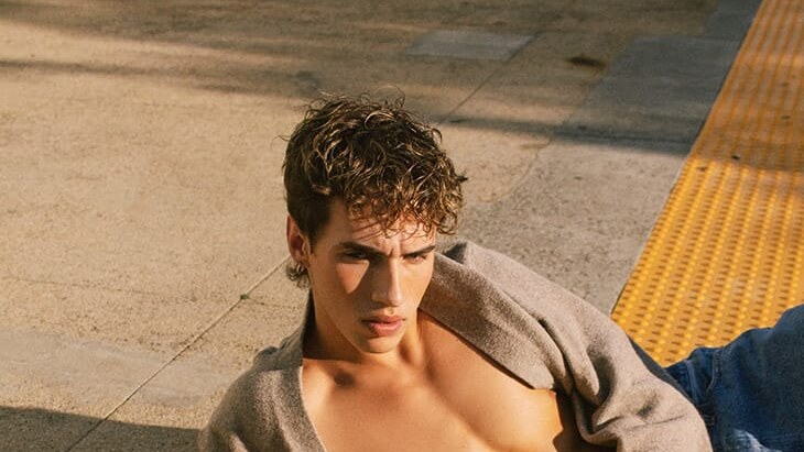 Jonah Folks reclines on a station platform in an open cardigan and jeans, with one hand resting on yellow tactile paving.