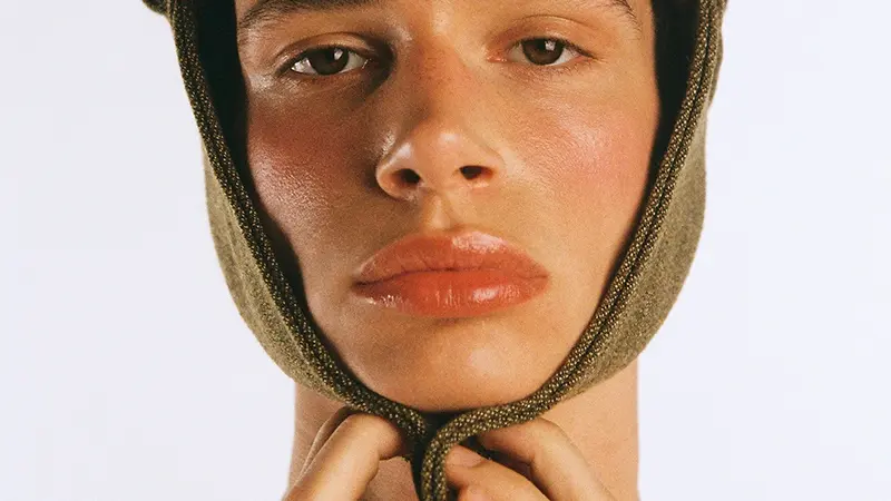 Close-up portrait of a model wearing a textured olive cap with ear flaps, holding the chin strap beneath their face.
