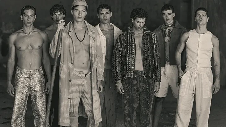 Seven male models stand together in varied menswear looks, including patterned trousers, layered tailoring, an open knit cardigan and relaxed light-colored separates.