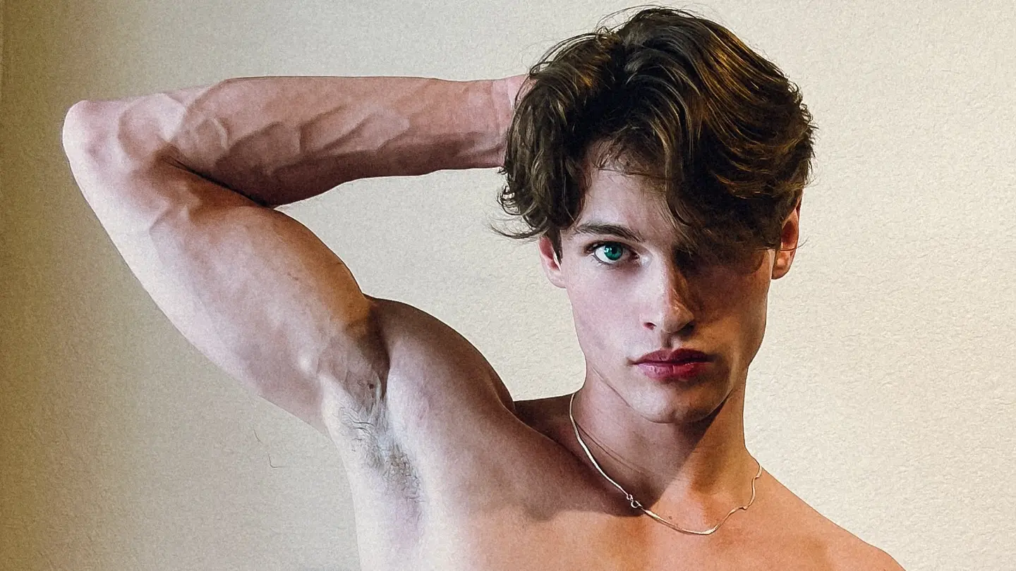 Dustin Ray Wilcher poses shirtless against a light wall, with one arm raised behind his head and a chain necklace visible.