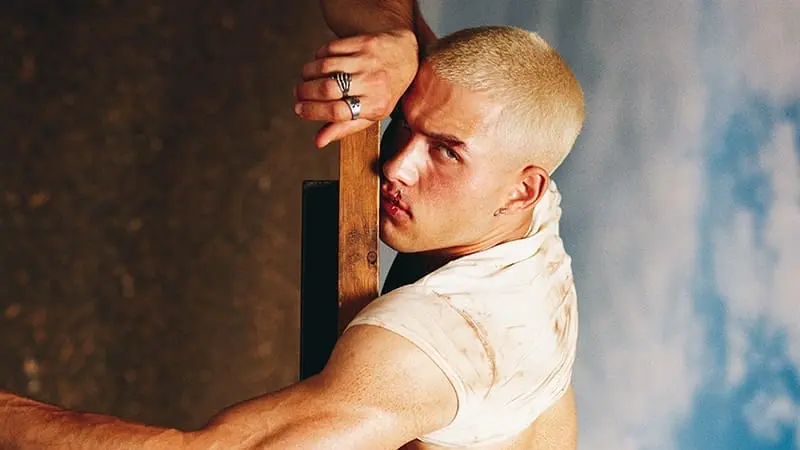 Lucas Sapoznick grips a wooden beam while looking back over his shoulder, wearing a cropped white top and jeans.