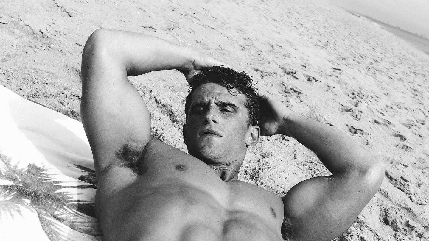 Matt van de Sande reclines shirtless on a sandy beach with his arms behind his head in a black-and-white portrait.