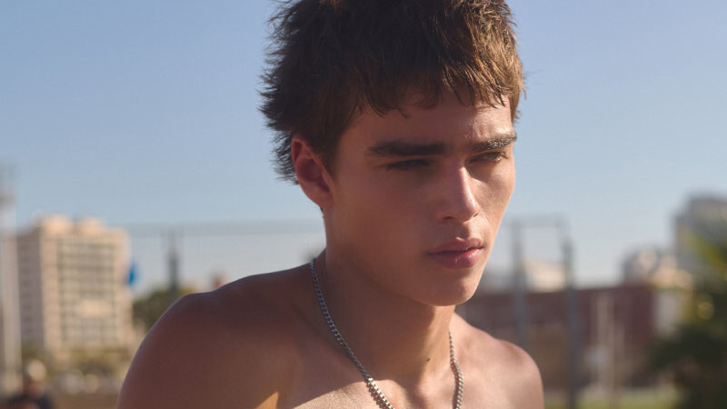 A young man holds a worn skateboard across his torso outdoors, wearing blue shorts and a chain necklace.