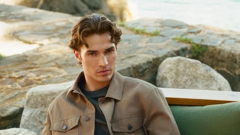 Callum Simpson sits outdoors on a green lounge chair among coastal rocks, wearing a taupe overshirt, dark top and white trousers.