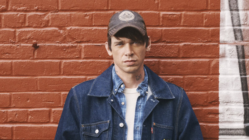 Cole Mohr stands against a red brick wall wearing a baseball cap, layered denim and jeans, with his hands in his jacket pockets.