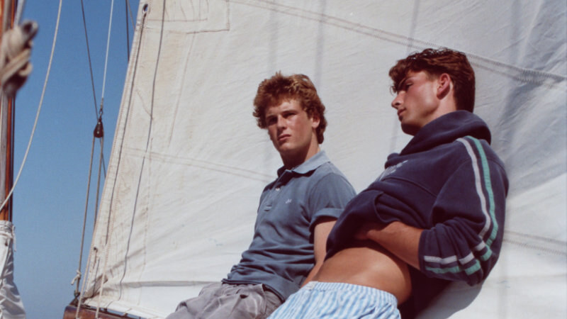 Two men in relaxed nautical clothing lean against a sail on a boat, with blue water visible behind them.