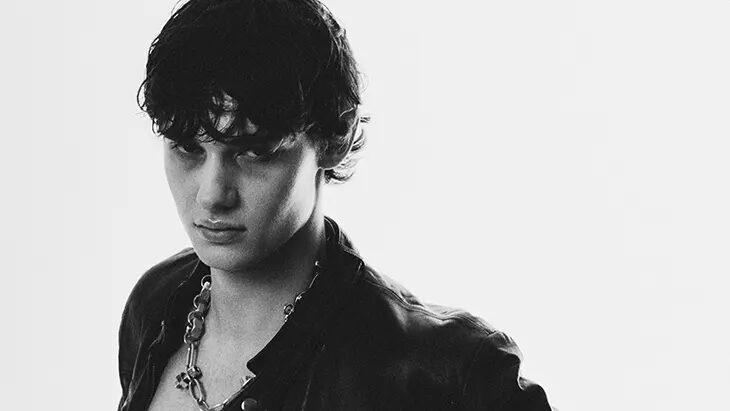 Juan Navarro poses in a black-and-white fashion portrait wearing an open leather jacket, chain necklace and dark trousers.