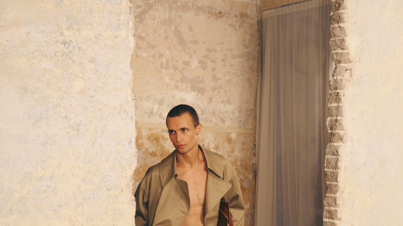 A male model steps through a doorway wearing a flowing tan coat, open shirtless torso, loose trousers and black shoes.