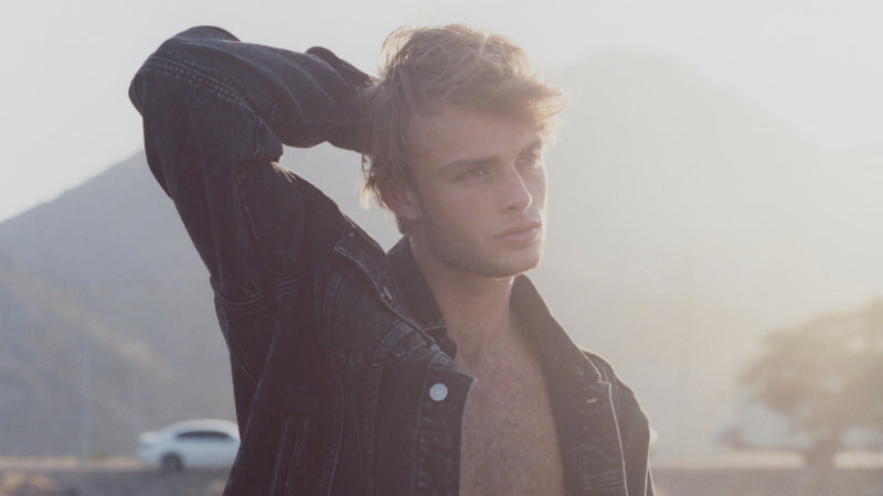 Model wearing an open dark denim jacket and red shorts stands on a beach in hazy sunlight, with a mountain and car in the background.