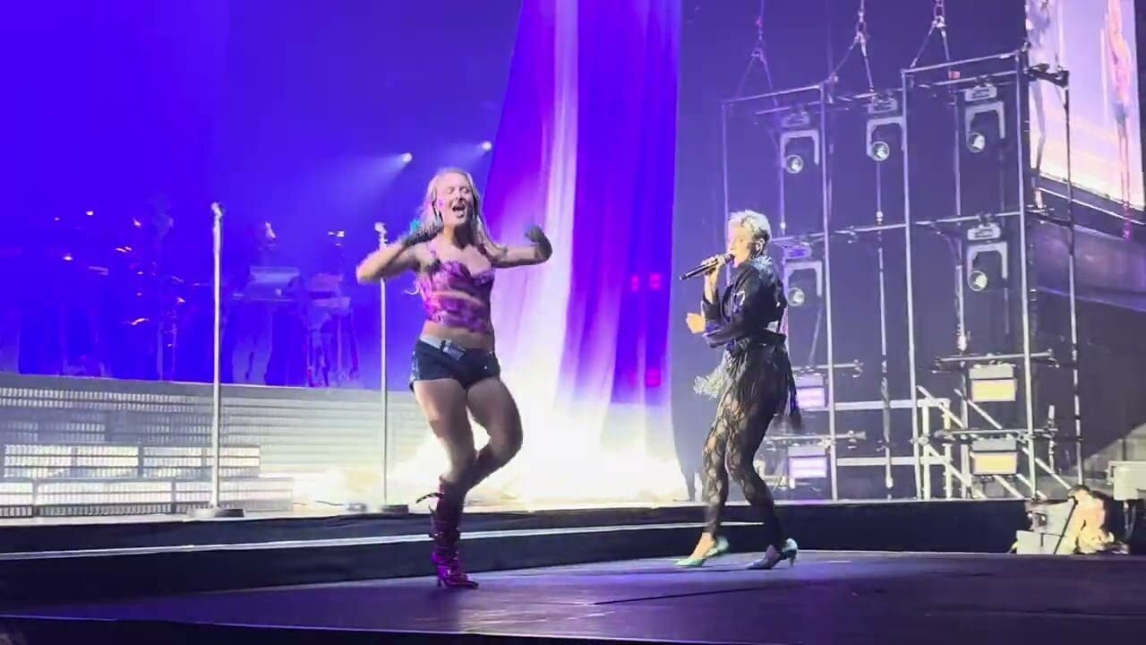 Two performers on a concert stage, one singing into a microphone while the other dances in front of purple lights