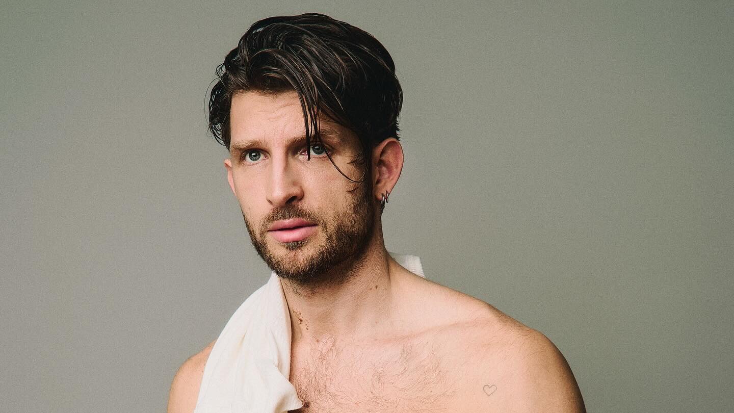 Konstantin Resch poses shirtless against a neutral gray background, with a white cloth over one shoulder, visible chest and body hair, and his hands clasped at his waist.