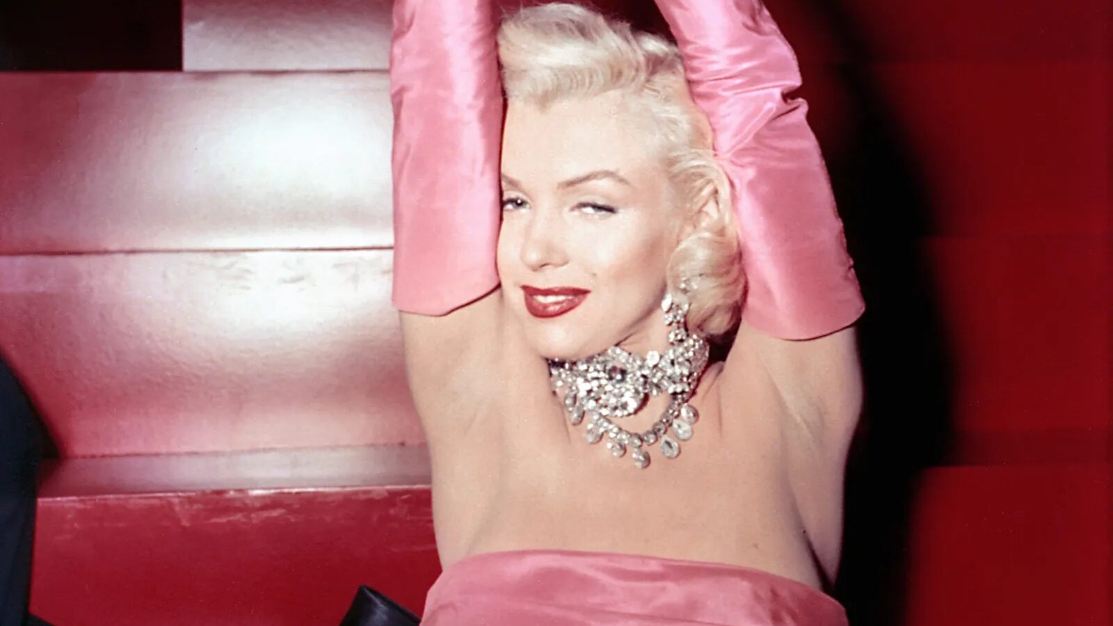 Marilyn Monroe sits on red steps wearing a pink satin gown, long gloves and sparkling jewelry, with both arms raised above her head.