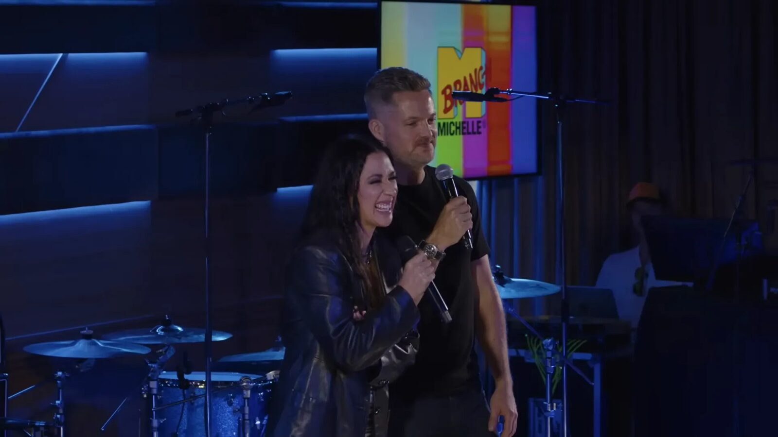 Michelle Branch and Damien Fahey hold microphones onstage during the MBTV special, with a colorful MBTV graphic behind them.