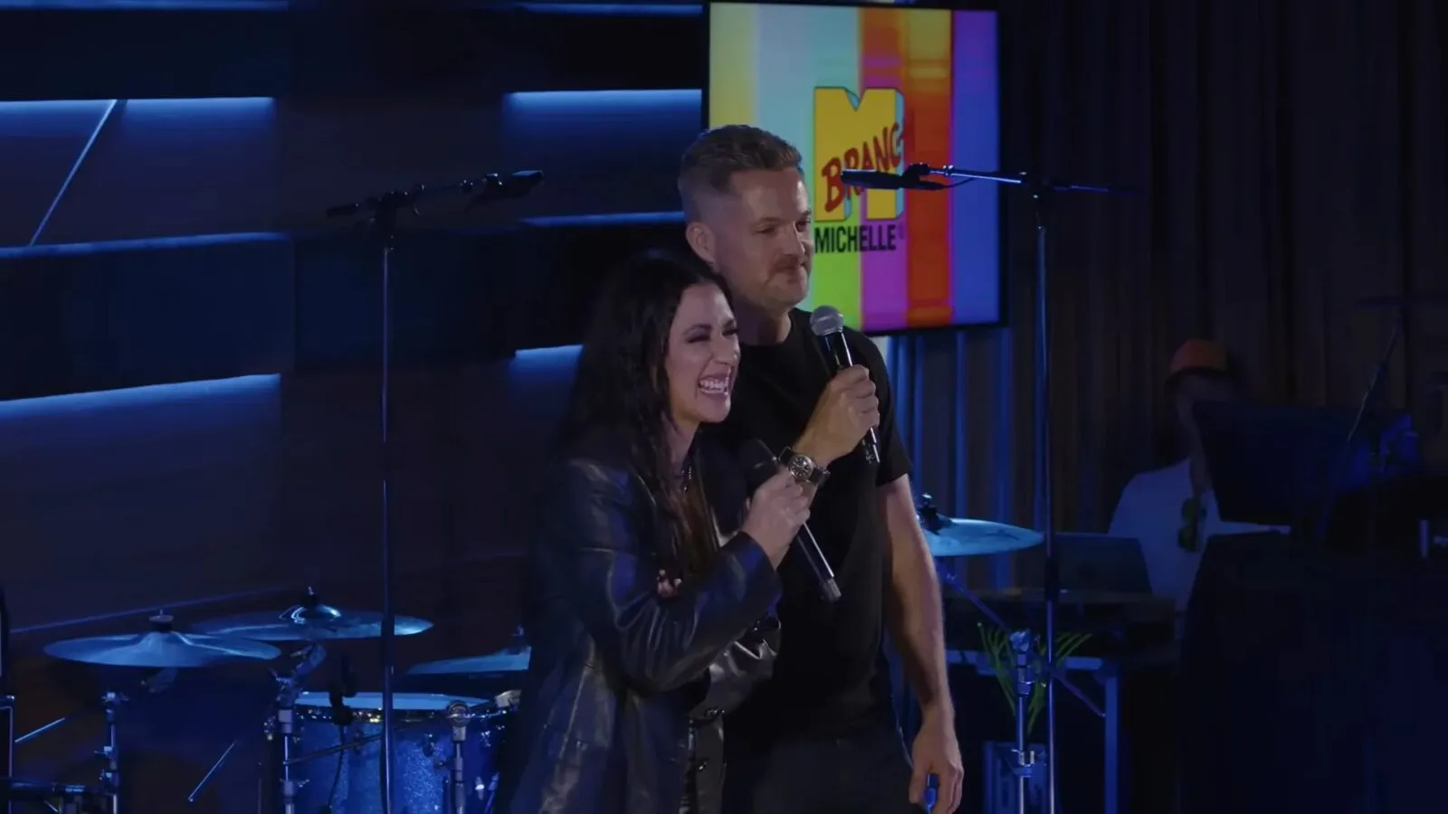 Michelle Branch and Damien Fahey hold microphones onstage during the MBTV special, with a colorful MBTV graphic behind them.