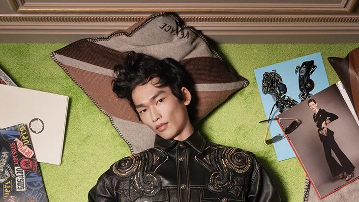 A male model lies on a green carpet surrounded by books and fashion accessories, wearing a black embellished leather jacket, blue trousers and black shoes.