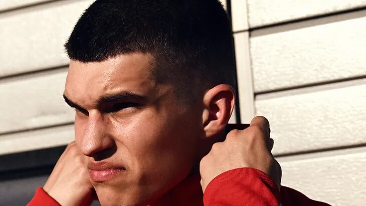Male model adjusting the collar of a red track jacket in a close-up portrait against a light-colored paneled wall.