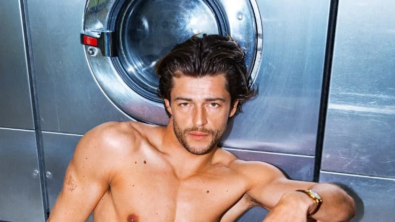 Sam Salter sits on a laundromat floor in grey boxer briefs and socks, leaning against a washing machine.