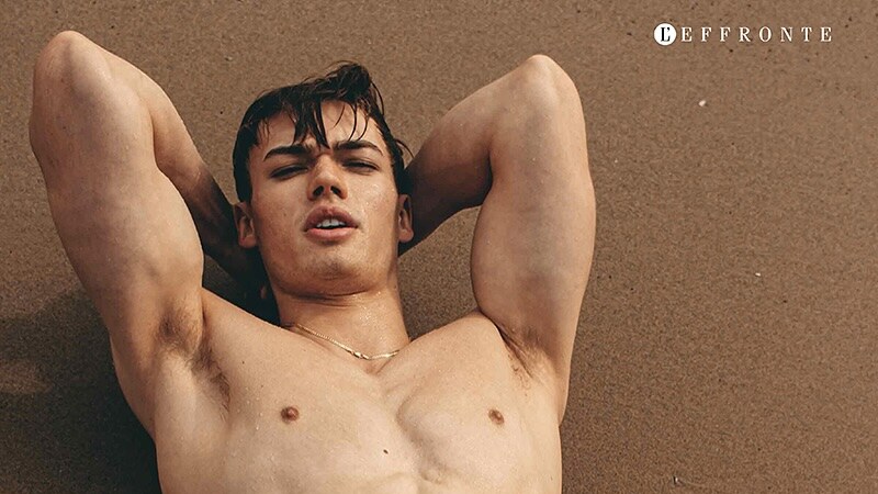 Colin Walling lies shirtless on a textured brown surface with his arms behind his head, water droplets visible on his skin.