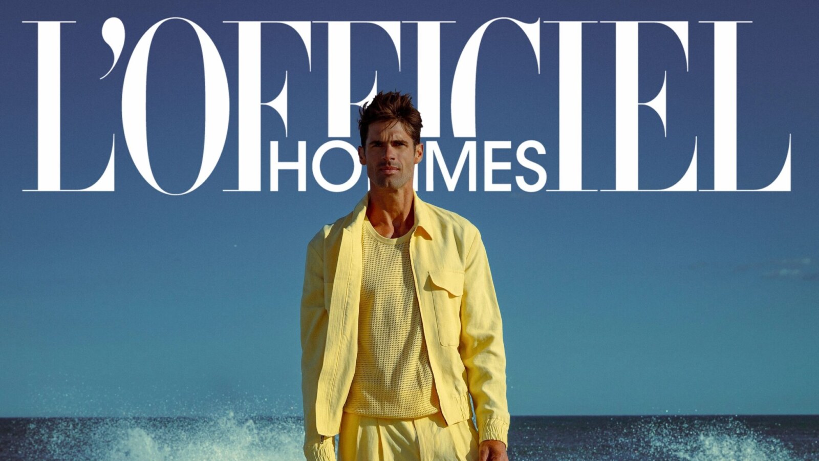 Chad White stands on a ladder in ocean surf wearing a yellow shirt, open jacket and trousers, with L’Officiel Hommes and his name displayed in large white type.