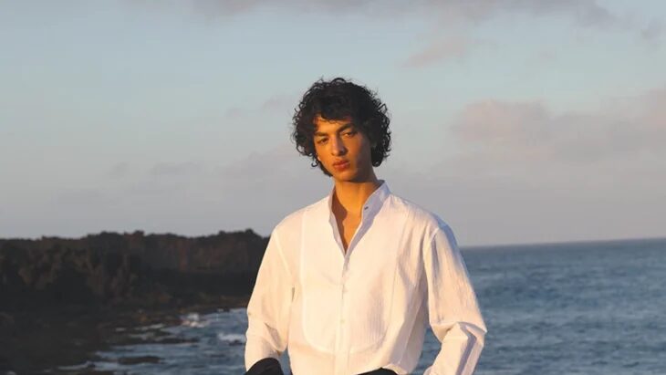 Teo Abihdana stands on a black volcanic shoreline in a white shirt and black trousers, holding a dark jacket with the ocean behind him.