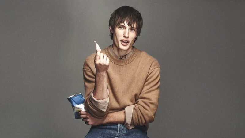 Thatcher Thornton sits on a stool wearing a camel crew-neck sweater, relaxed blue denim and brown shoes, holding a small blue package and a pale object against a gray studio backdrop.