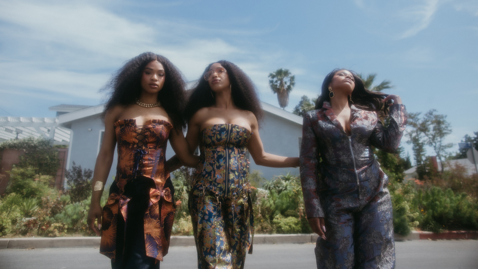 The Womack Sisters stand together outdoors in coordinated patterned outfits, with two facing the camera and one looking upward.