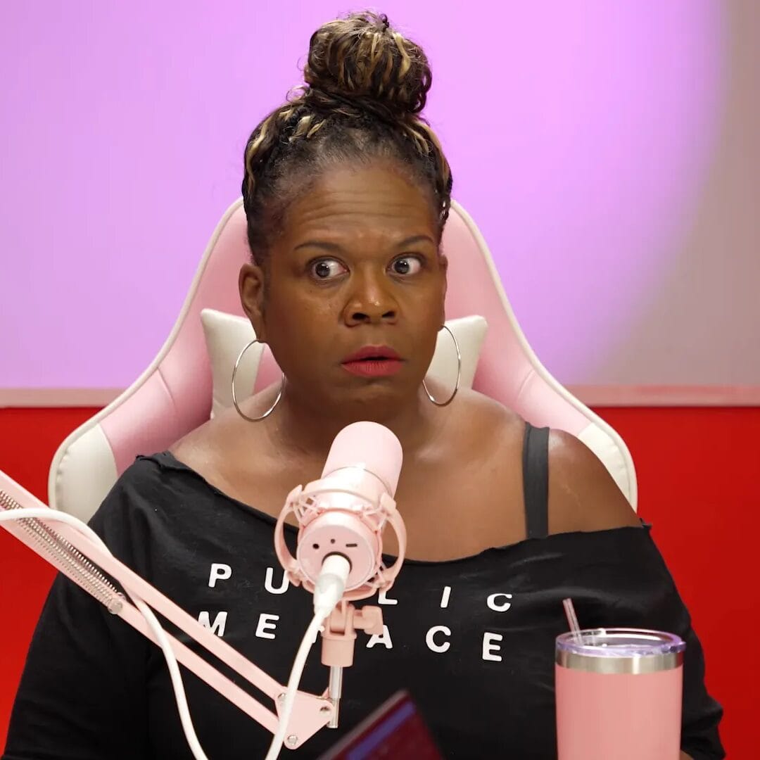 Yamaneika Saunders sits behind a pink microphone in a podcast studio, looking toward the camera with a surprised expression.
