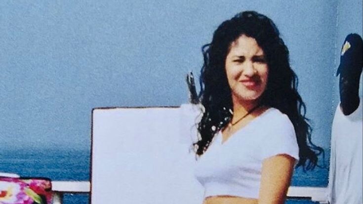 Selena Quintanilla smiles while standing in a white crop top and floral pants near a waterside railing.