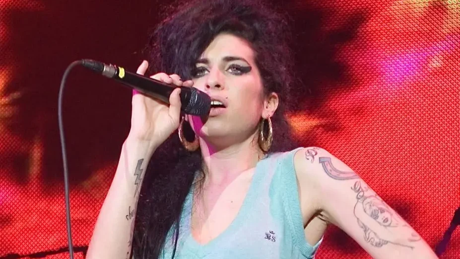 Amy Winehouse sings into a handheld microphone onstage, wearing a light blue sleeveless top with one hand on her hip.