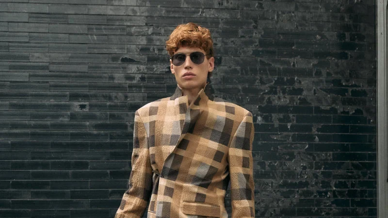 Model wearing a full checked brown, beige and gray suit with sunglasses and carrying a black bag against a dark brick wall.