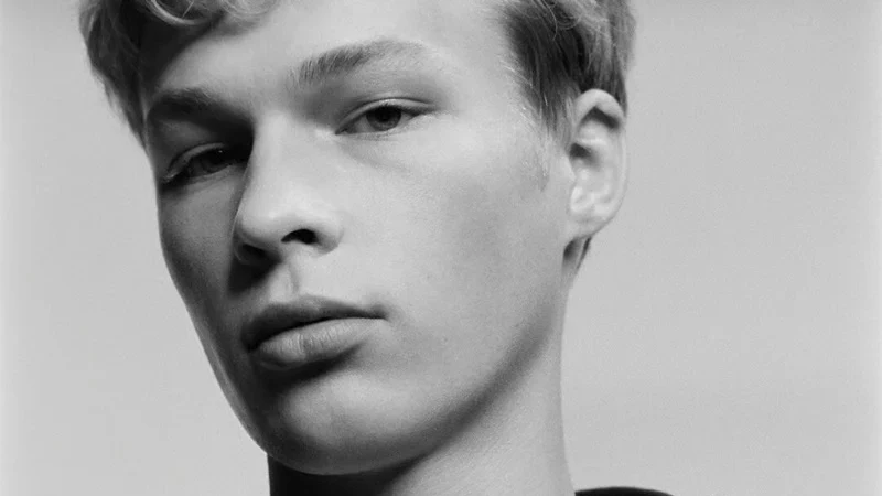 Black-and-white close portrait of a male model wearing a white collared shirt, striped Dior tie and dark zip-front jacket.