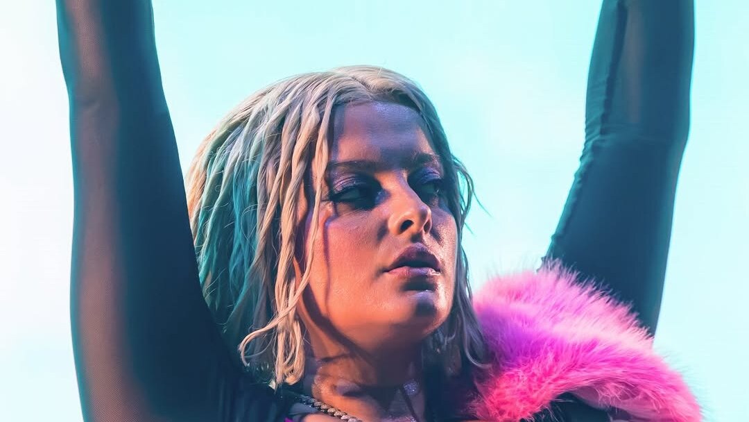 Bebe Rexha performs with both arms raised, holding a microphone against a bright blue stage backdrop.