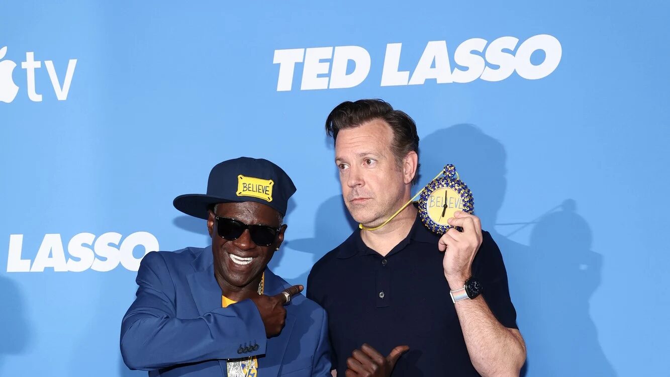 Flavor Flav and Jason Sudeikis pose together, with Sudeikis holding a custom yellow “BELIEVE” clock necklace.