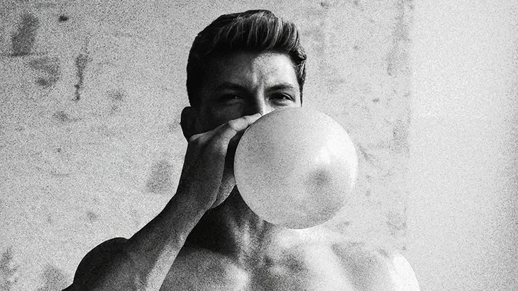 Mate sits shirtless on a chair, holding a pale balloon in front of his mouth in a grainy black-and-white portrait.