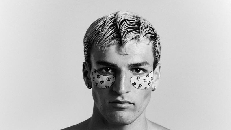 Milhan Bouzidi faces the camera in a high-contrast black-and-white portrait, wearing patterned eye patches and small hoop earrings.