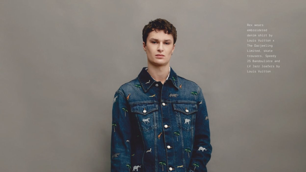 Rex Rayner stands against a gray backdrop in an embroidered denim jacket and matching trousers, holding a patterned handbag.