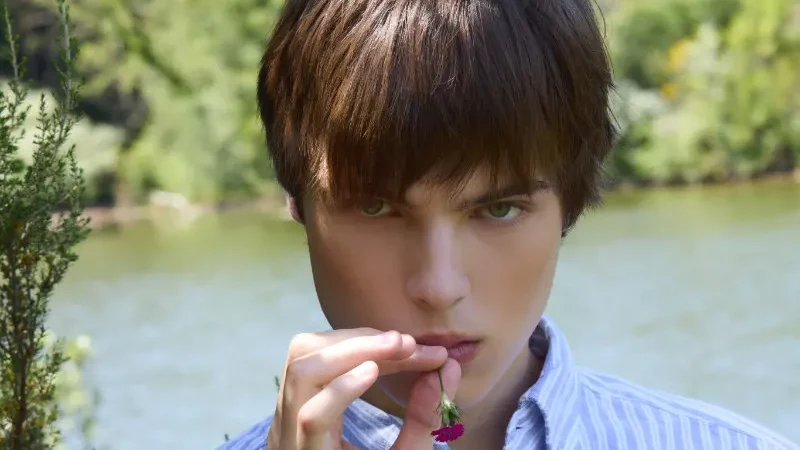 A young male model in a blue-and-white striped shirt holds a small flower near his lips beside a lake.