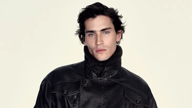 Male model wearing a belted black leather jacket and dark trousers against a pale background.