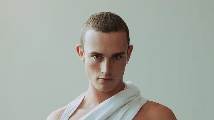 Harry Ash faces the camera in a white draped top and black jeans against a pale background.