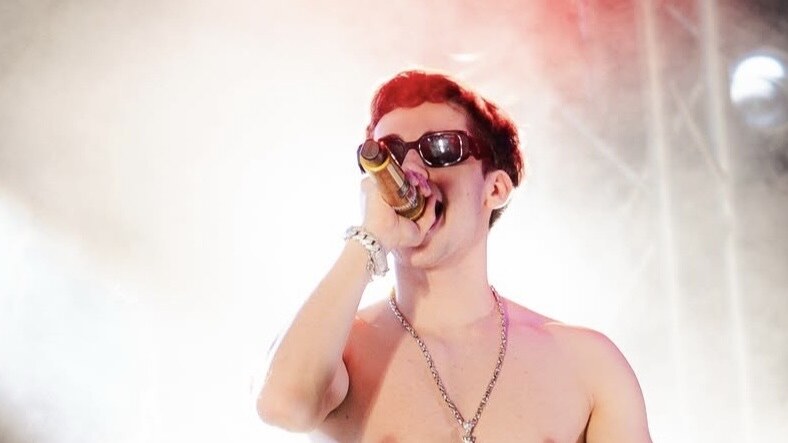 Kevin AMF performs shirtless onstage while wearing sunglasses and holding a microphone amid bright lights and haze.