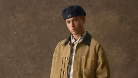 A model wears a tan jacket over a plaid shirt with loose trousers, black boots and a beret while carrying a structured bag.