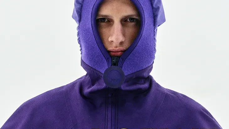 Model wearing a purple hooded LOEWE jacket with bright yellow lettering and an exposed midriff against a pale background.