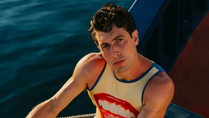 Carlos Lara Herrera poses on a boat above dark water, wearing a graphic tank top and red briefs while holding a metal wheel.