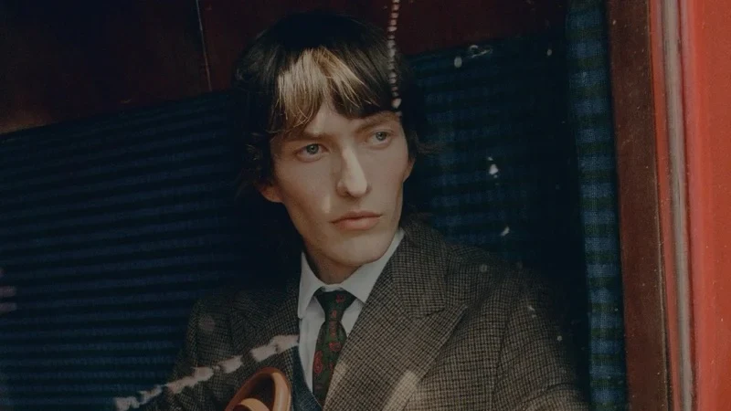 Erik Kettschick sits inside a train carriage wearing a checked suit and tie while holding a brown leather travel bag.