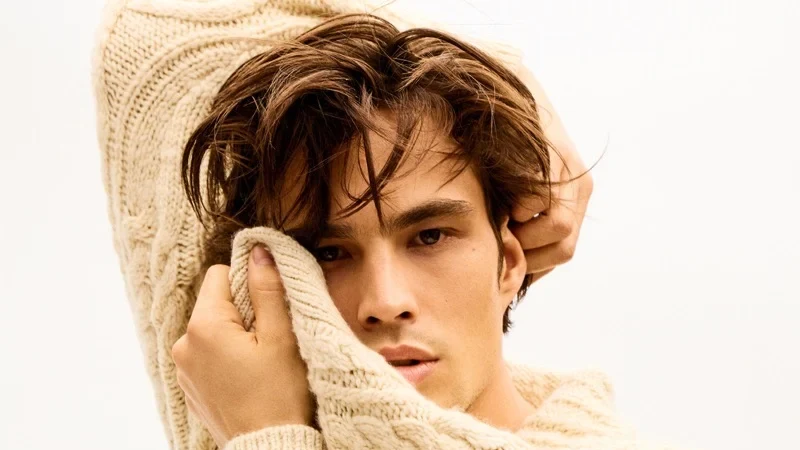 Louis Baines poses in a cream cable-knit sweater, with one sleeve raised behind his head and another hand touching his face.
