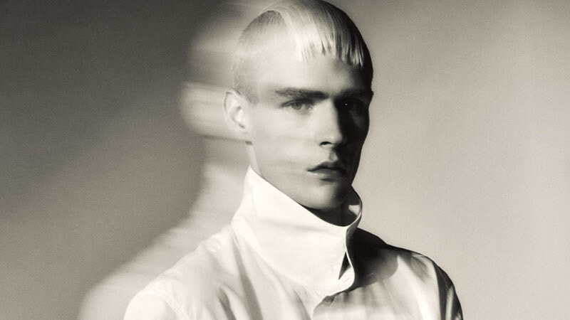 Paolo Grebic poses in a high-collared white shirt while holding a large quilted white cushion, rendered in a softly blurred monochrome fashion portrait.