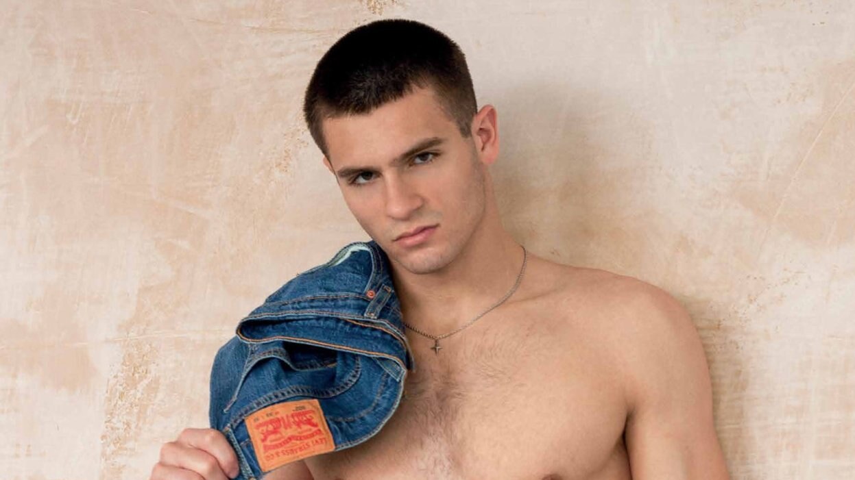 Marcello Maffezzoli poses shirtless against a textured wall, holding a denim jacket over one shoulder and wearing white Emporio Armani briefs.